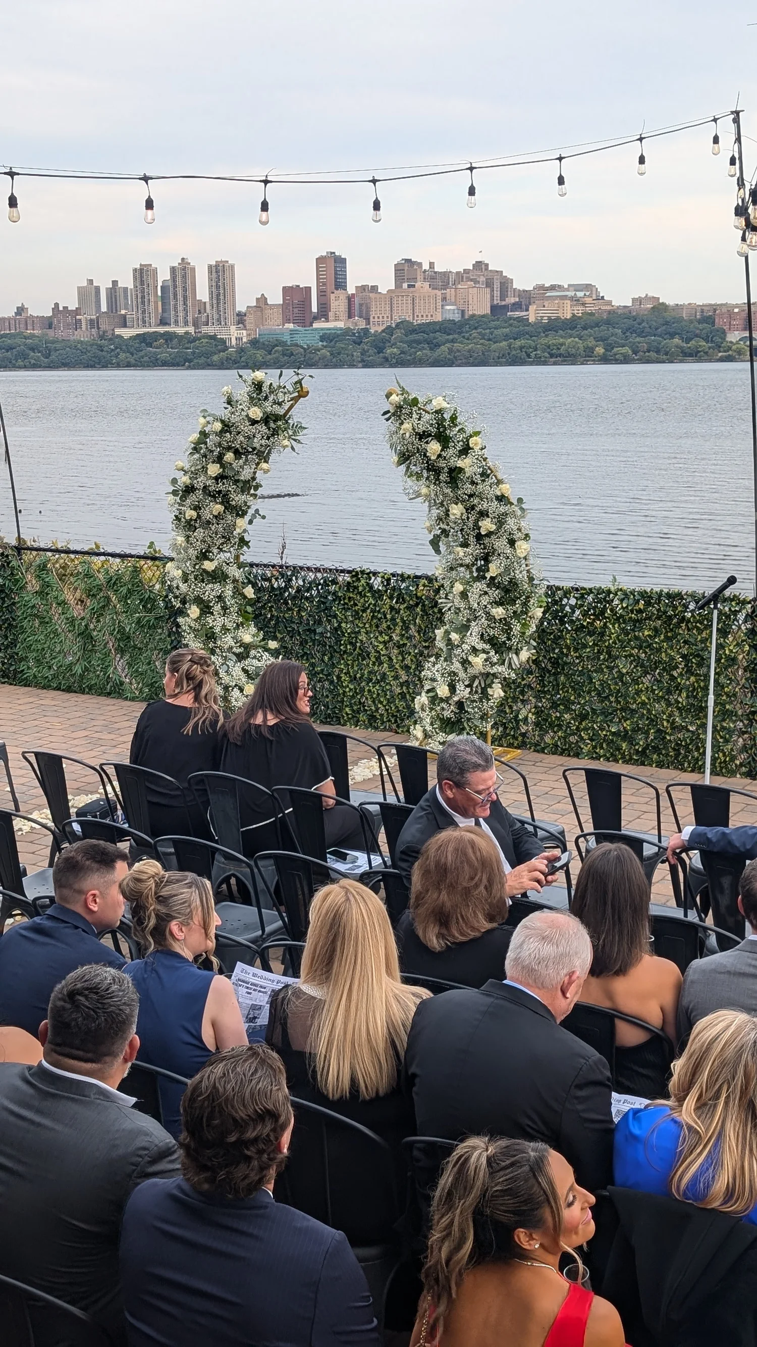 A wedding ceremony on the DeNovo Edgewater patio with a floral arch and the Manhattan skyline across the Hudson.