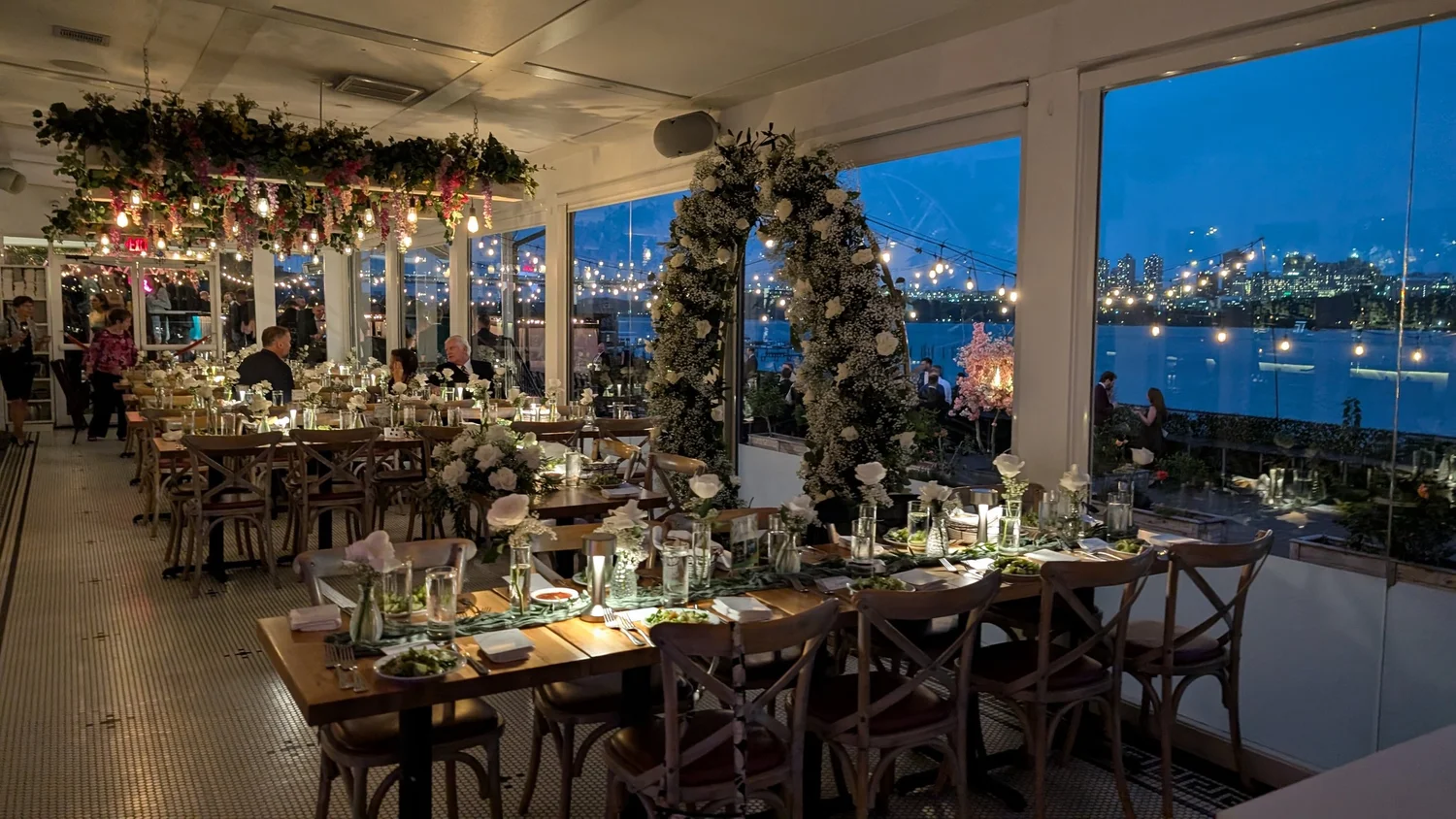 A wedding reception at dusk with long tables, florals, and the lit Manhattan skyline across the river.