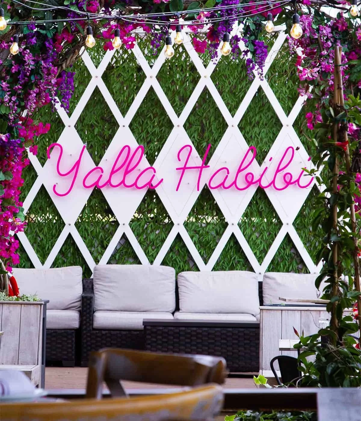 The DeNovo Edgewater patio lounge corner with a neon sign, florals, and rattan seating.