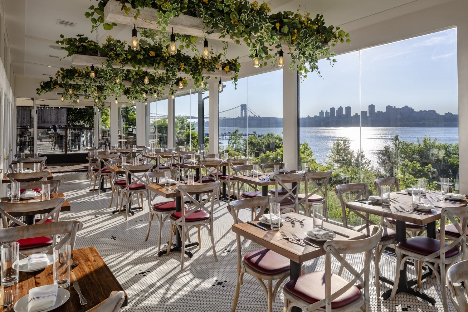 The DeNovo Edgewater glass-walled sunroom dining area framing the Hudson River and George Washington Bridge.
